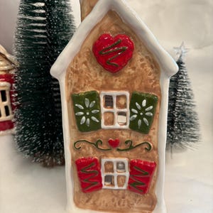 May include: A ceramic gingerbread house with a brown body, white roof, and chimney. It features a red heart, green shutters, and red window decorations. The house is decorated with white icing and sits among miniature Christmas trees.