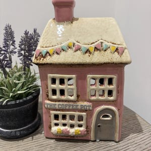 Peut inclure: Un bougeoir en céramique en forme de maison rose et crème avec une cheminée, portant l'inscription THE COFFEE POT au-dessus de la fenêtre du rez-de-chaussée. Le toit est orné de guirlandes colorées et la façade présente des fenêtres et une porte.