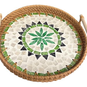 Puede incluir: Bandeja redonda de servicio de ratán tejido con un diseño de mosaico. El mosaico presenta un patrón central de hojas verdes, rodeado de azulejos verdes, negros y blancos. La bandeja tiene dos asas de madera.