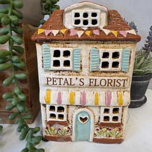 May include: A ceramic house-shaped ornament with the text "PETAL'S FLORIST". The house features a brown tiled roof, blue shutters, and a light blue door with a heart-shaped cutout. The awning is striped with yellow and pink. The figurine is decorated with colourful details.