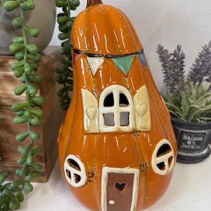 Puede incluir: Casa de cerámica naranja con forma de calabaza y tallo marrón. La casa tiene una puerta con un recorte de corazón, ventanas con contraventanas y una pancarta decorativa. Es un objeto decorativo.