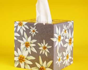 May include: A square tissue box with a gray background and a repeating pattern of white and yellow flowers. White tissues are pulled from the top. The background is a bright yellow.