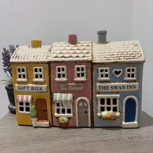 May include: A ceramic row house figurine featuring three connected buildings in yellow, pink, and blue. The yellow building is labeled GIFT BOX, the pink building is labeled Tea Rooms, and the blue building is labeled THE SWAN INN with a heart icon.