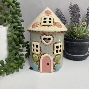 Może przedstawiać: Ceramiczny świecznik w kształcie domu z jasnoniebieskim korpusem, kremowym dachem i różowymi kwiatowymi akcentami. Dom posiada okno w kształcie serca, różowe drzwi i kwadratowe okna. Małe drzewka w donicach flankują drzwi.