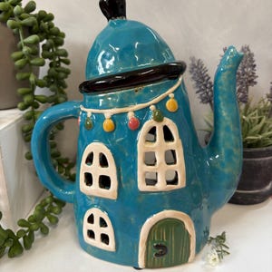 May include: A teal ceramic teapot shaped like a house, with white-framed windows and a green door. The teapot has a black handle and spout, with colourful accents around the roofline. A decorative piece for the home.