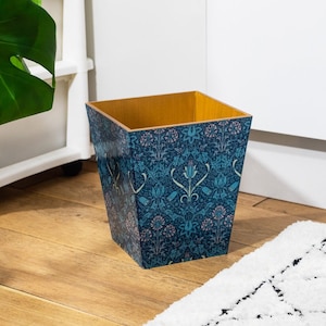 Puede incluir: Una papelera cuadrada azul y dorada con un patrón floral. El patrón presenta flores y hojas estilizadas en tonos de azul, rosa y verde.