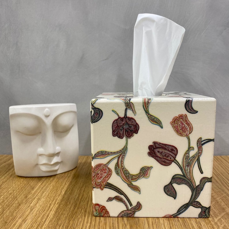 May include: White ceramic tissue box cover with a floral design featuring red, pink, and green tulips on a cream background. The tissue box cover has a white tissue box inside.