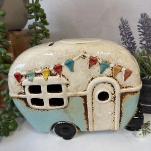 Può includere: Figurina di camper in ceramica con base azzurra e parte superiore bianco sporco. Il camper presenta una porta, una finestra e bandierine colorate. Le ruote sono nere e il design ha un aspetto rustico e artigianale.