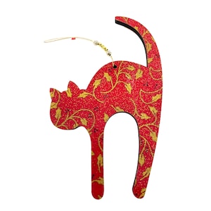 May include: A red cat-shaped ornament with a gold floral pattern. The ornament has a black outline and a gold string for hanging. The cat is in a standing pose with its tail curved upwards. The ornament is likely made of wood.