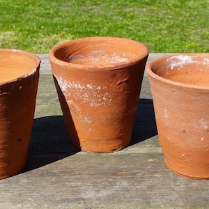 May include: Three terracotta plant pots of different sizes on a wooden surface. The pots are a warm reddish-brown colour, with a slightly rough texture. The pots are empty and ready for planting.