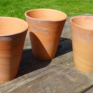 May include: Three terracotta plant pots with a conical design, set on a weathered wooden surface. The pots are unglazed, showing a natural reddish-brown colour. Green grass forms the background, indicating an outdoor location.