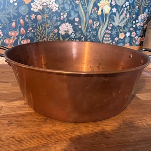 May include: A large copper pot with black handles. The pot has a wide, rounded shape and a shiny surface. It sits on a wooden surface, with floral wallpaper in the background. The pot is likely for cooking.