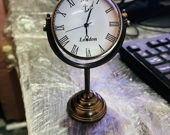 May include: A vintage-style clock with a white face, black Roman numerals, and the word "London" printed at the bottom. The clock is set in a bronze-colored metal frame and stand. The clock's hands point to approximately 1:35.