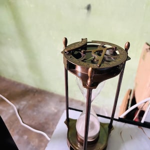 Puede incluir: Un reloj de arena antiguo de latón con diseño de reloj de sol y brújula. El reloj de arena tiene un centro de vidrio transparente lleno de arena blanca, sostenido por un marco de latón decorativo. El reloj de sol presenta números romanos y una aguja de brújula.