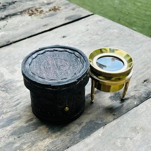 May include: A vintage brass magnifying glass with a round lens and three small legs, next to a dark brown leather case with a decorative top. The case has a gold-colored stud. The items are on a weathered wooden surface.