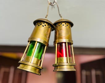 Op de afbeelding: Twee antieke messing lantaarns die aan een plafondhaak hangen. Eén lantaarn heeft een groene glazen lens, de andere een rode lens. De lantaarns hebben een cilindrische vorm met decoratieve perforaties en een klein handvat.