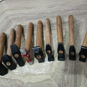 Puede incluir: Una colección de martillos con mangos de madera y cabezas negras o plateadas. Los martillos están dispuestos en fila, mostrando sus diferentes tamaños y formas. Los mangos de madera tienen un acabado natural.