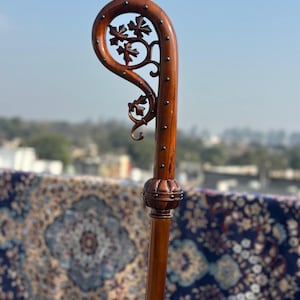 May include: A brown wooden walking stick with a curved handle and floral detailing. The handle has a carved floral design and is studded with small metal pins. The stick has a decorative bulbous section. The wood has a polished finish, and the stick is a rich brown colour.