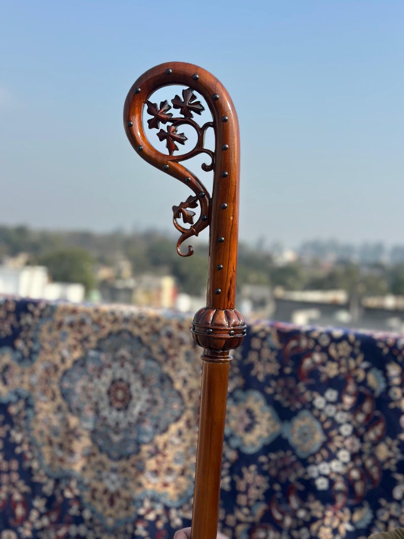 May include: A brown wooden walking stick with a curved handle and floral detailing. The handle has a carved floral design and is studded with small metal pins. The stick has a decorative bulbous section. The wood has a polished finish, and the stick is a rich brown colour.