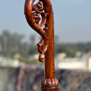 May include: A wooden walking stick with a curved handle and floral carvings. The dark brown wood is studded with small, dark metal pins. The base has a decorative, rounded design. The overall height is approximately 100 cm.
