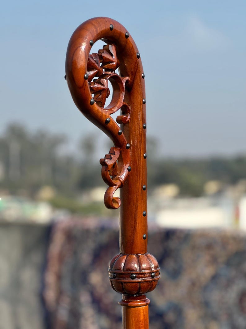 May include: A wooden walking stick with a curved handle and floral carvings. The dark brown wood is studded with small, dark metal pins. The base has a decorative, rounded design. The overall height is approximately 100 cm.