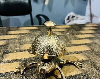 May include: A brass frog-shaped bell with a domed top and a small handle. The bell has intricate leaf-like patterns and is set on a striped surface. The frog's legs are outstretched, giving it a playful appearance.