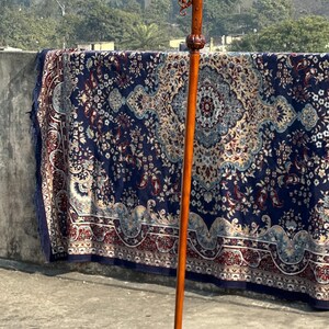 May include: A long, wooden walking stick with a curved handle. The handle has detailed floral carvings. The stick is a warm, reddish-brown colour and stands upright. The handle has a decorative design.