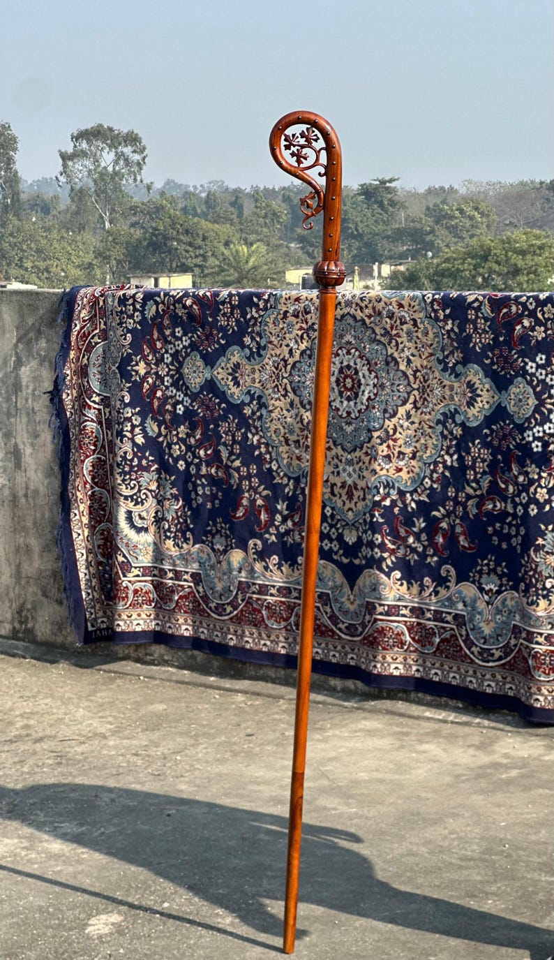 May include: A long, wooden walking stick with a curved handle. The handle has detailed floral carvings. The stick is a warm, reddish-brown colour and stands upright. The handle has a decorative design.
