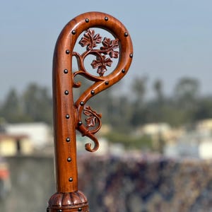May include: A handcrafted wooden walking stick, featuring a curved handle with detailed leaf carvings. The dark brown wood is adorned with small metal studs. The handle showcases a decorative floral design, adding a touch of elegance to the cane.