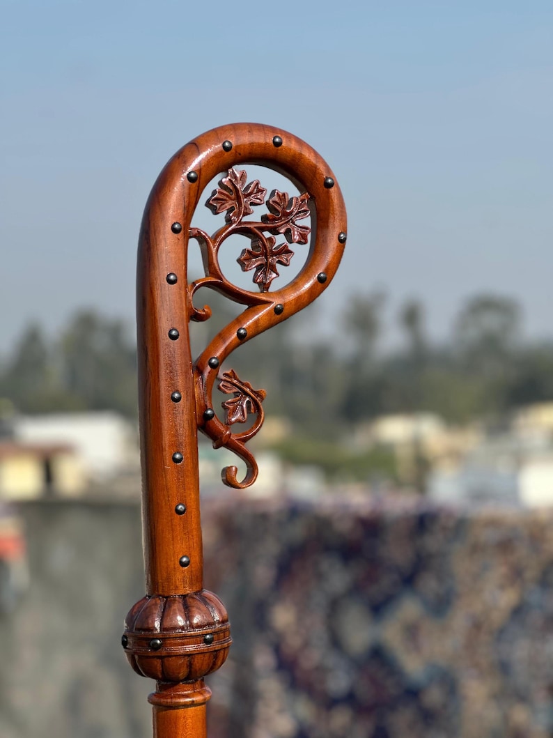 May include: A handcrafted wooden walking stick, featuring a curved handle with detailed leaf carvings. The dark brown wood is adorned with small metal studs. The handle showcases a decorative floral design, adding a touch of elegance to the cane.