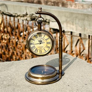 May include: A vintage brass desk clock with a compass base. The clock face is cream-colored with Roman numerals and the text "VICTORIA LONDON 1915". The clock is supported by a curved brass arm and sits on a circular base with a compass.