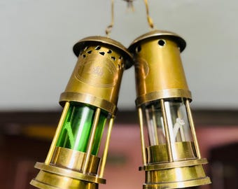 Op de afbeelding: Twee antieke messing olielampen die aan een haak hangen. Eén lamp heeft een groene glazen binnenkant, de andere is helder. Beide lampen hebben een cilindrische vorm met een decoratieve boven- en onderkant en worden opgehangen aan een messing ketting.