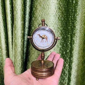 Op de afbeelding: Een vintage messing klok met een ronde wijzerplaat met een kameelillustratie. De klok heeft Arabische cijfers en wordt ondersteund door een gelaagde basis. De klok wordt in een hand gehouden tegen een groen patroon.