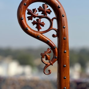 May include: A wooden walking stick with a curved top and a decorative floral design. The dark brown wood is accented with small, dark metal studs. The floral design features carved leaves and vines.