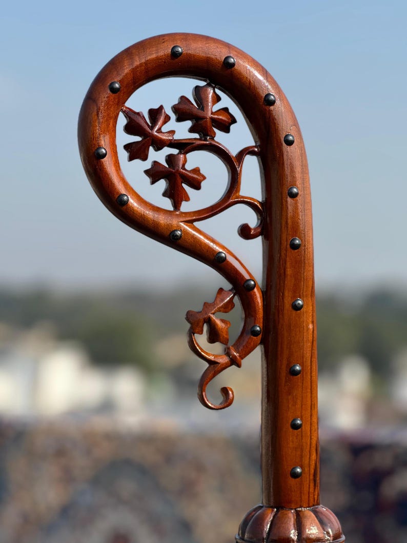 May include: A wooden walking stick with a curved top and a decorative floral design. The dark brown wood is accented with small, dark metal studs. The floral design features carved leaves and vines.