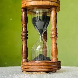 May include: An hourglass featuring a dark brown wooden frame with turned pillars connecting circular top and bottom bases. The clear glass bulb contains fine black sand, with a portion of the sand currently resting in the bottom chamber.