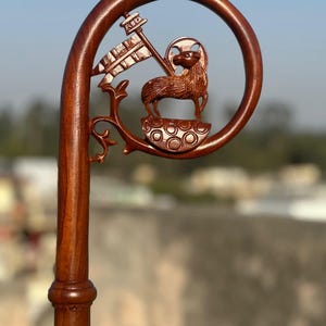 May include: A carved wooden staff with a curved top featuring a lamb holding a flag. The staff is a rich brown color and has intricate details, including a circular design around the lamb.