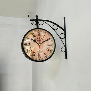 Pode incluir: Relógio de parede de dupla face com estrutura de metal preta e mostrador em estilo vintage. O mostrador do relógio apresenta algarismos romanos e o texto "VICTORIA STATION 1747 LONDON". O relógio está montado numa parede branca.