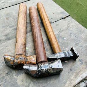 Puede incluir: Tres martillos con mangos de madera y cabezas de metal descansan sobre una superficie de madera envejecida. Los mangos varían en color de marrón claro a oscuro. Las cabezas de los martillos muestran signos de desgaste y óxido.