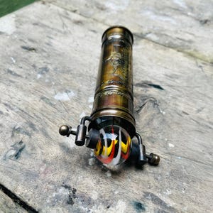 May include: A vintage brass kaleidoscope with a decorative etched design. The kaleidoscope features a colorful glass ball with red, yellow, and white swirls. The instrument is set against a weathered wooden surface.