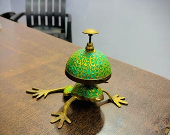 May include: A brass frog-shaped bell with a green and gold patterned dome. The bell has outstretched legs and a small handle on top. The bell is sitting on a wooden surface.