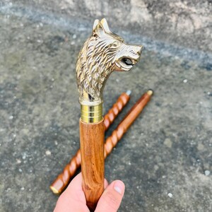 Puede incluir: Bastón con un mango de cabeza de lobo de latón y un eje de madera. La cabeza de lobo está detallada con pelaje texturizado y boca abierta. El eje es de color marrón claro. Dos bastones adicionales están en el fondo.