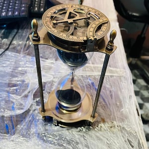 Puede incluir: Un reloj de arena de latón con un reloj de sol en la parte superior. El reloj de arena tiene una bombilla de vidrio transparente llena de arena negra. El reloj de sol presenta números romanos y un diseño de brújula. El marco está sostenido por cuatro postes de latón.