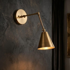 May include: A wall-mounted swing arm lamp with a brushed brass finish. The fixture features a circular wall plate, a two-part adjustable arm with visible joints, and a conical shade that directs light downward, creating a warm glow.