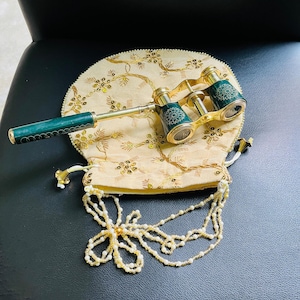 May include: An antique opera glass with a green handle and gold accents, resting on a cream-colored, embroidered pouch. The pouch has a beaded drawstring and a beaded flower design.