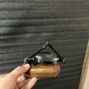 May include: A vintage magnifying glass with a wooden base and adjustable metal frame. The magnifying glass has a circular lens and is designed for reading or detailed inspection. The wooden base is a warm brown colour.