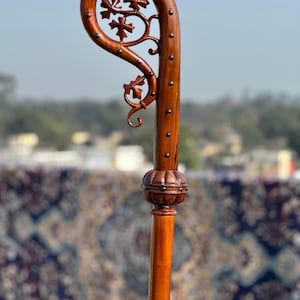 May include: A wooden shepherd's crook, stained a rich brown, with a curved top adorned with carved floral patterns and metal studs. The staff has a detailed, bulbous section. The design suggests a handcrafted, traditional item, likely used for ceremonial purposes.