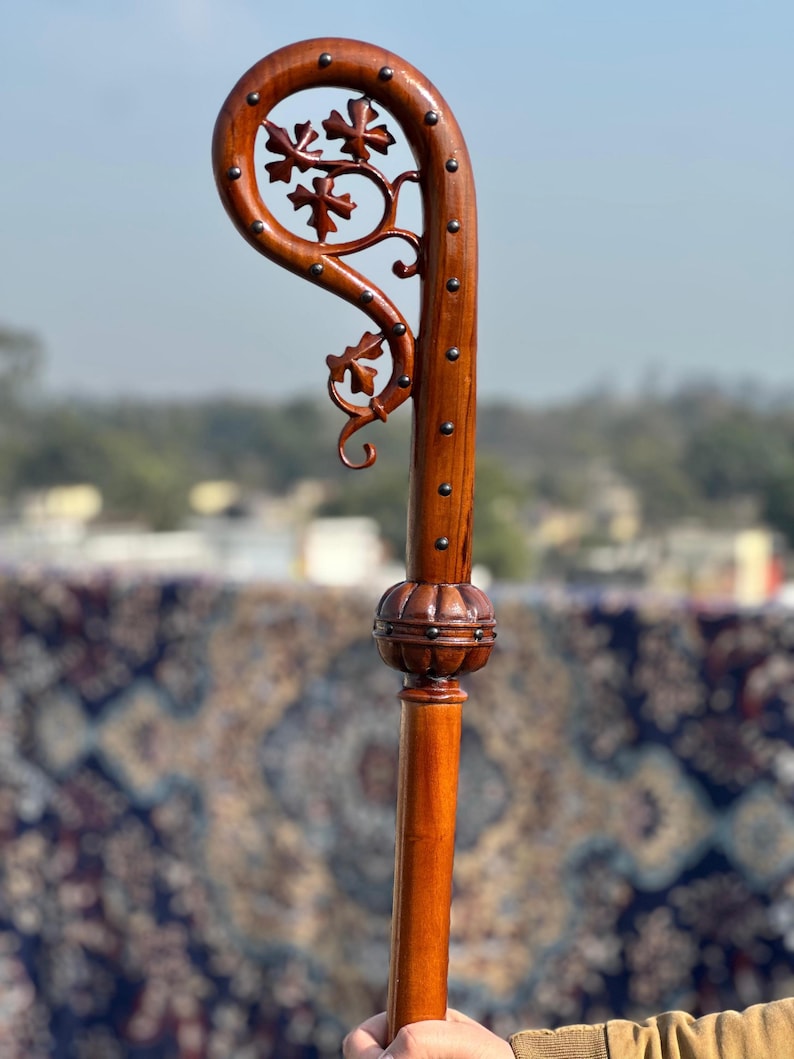 May include: A wooden shepherd's crook, stained a rich brown, with a curved top adorned with carved floral patterns and metal studs. The staff has a detailed, bulbous section. The design suggests a handcrafted, traditional item, likely used for ceremonial purposes.
