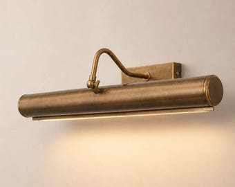 May include: A wall-mounted picture light with an antique brass finish. The fixture features a horizontal cylindrical lamp head attached to a rectangular wall plate by a curved, adjustable arm, emitting a warm downward glow of light.