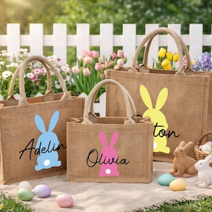 Könnte beinhalten: Drei Tragetaschen aus Jute mit Hasensilhouetten in Blau, Rosa und Gelb. Auf den Taschen sind Namen aufgedruckt. Osterei-Dekorationen umfassen einen Eierkorb und Hasenfiguren.